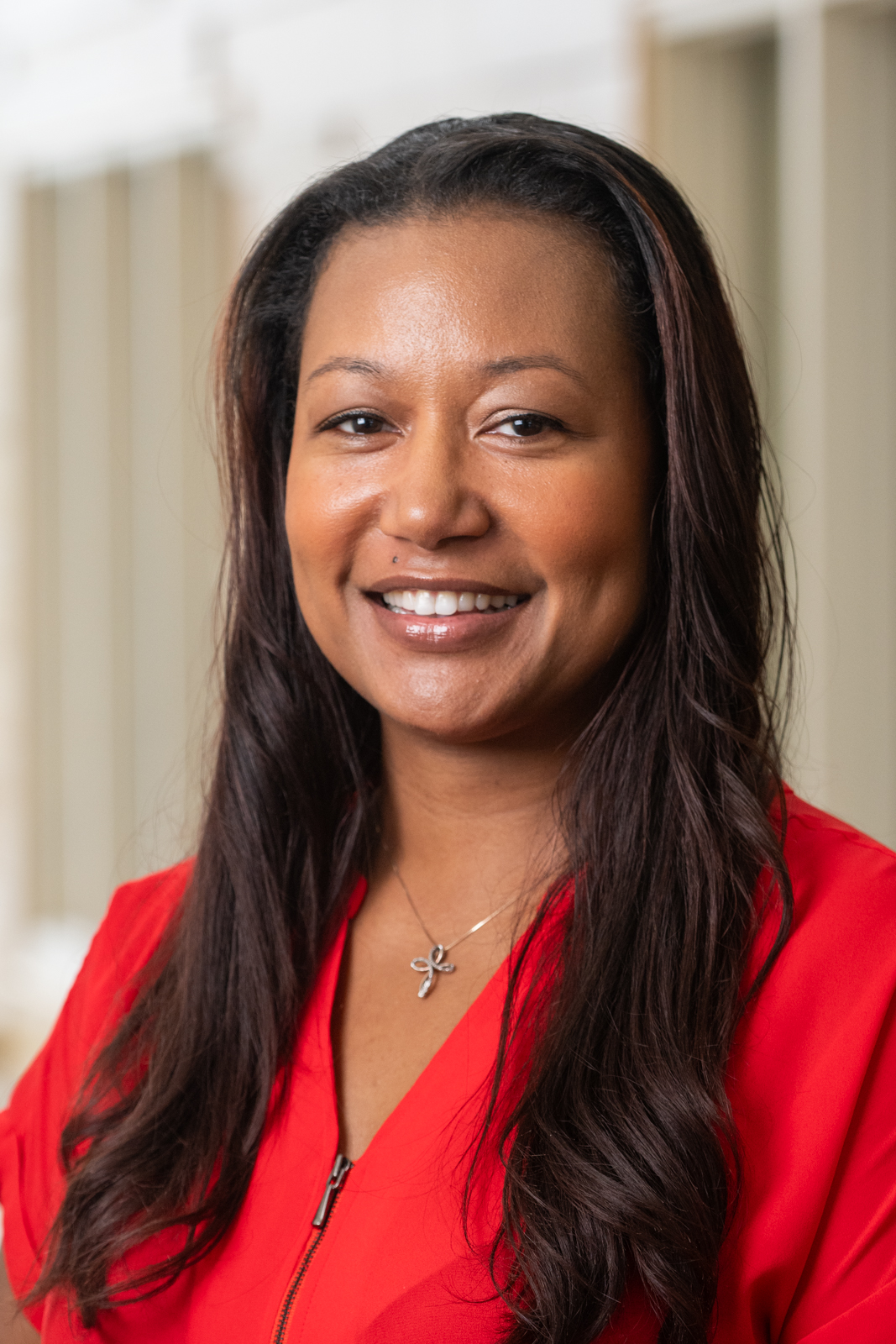 A portrait of Shaunte Rhodes. Shaunte is a caramel skin tone African-American woman in a red shirt with long black wavy hair. Shaunte is wearing a simple cross necklace and a cheerful smile.