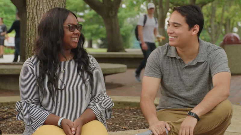 Two students dressed in fall clothing laughing with trees in the background