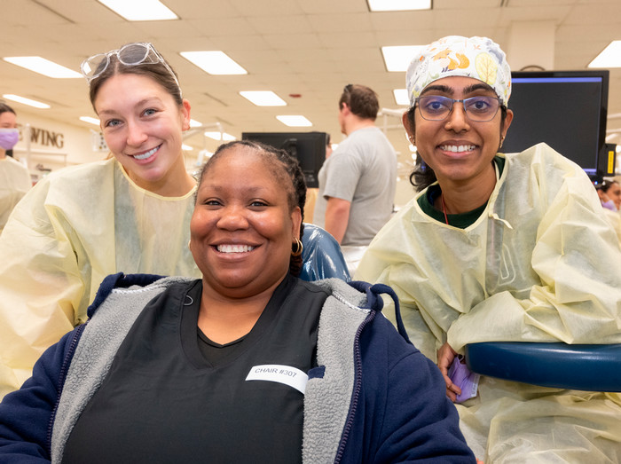 two female dental students in pale yellow scrubs pausing to take a photo with their patient a woman veteran in the dental chair seated between them